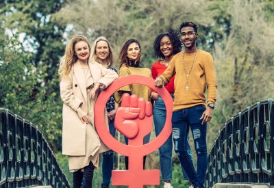 5 multiethnic people around a big cork feminist symbol smiling and looking at the camera. Feminism